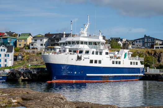 View of the Teistin ferry docked in a harbor with colorful houses and rocky terrain in the background in the Faroe Islands. View of the Teistin ferry docked in a harbor with colorful houses and rocky terrain in the background in the Faroe Islands.
