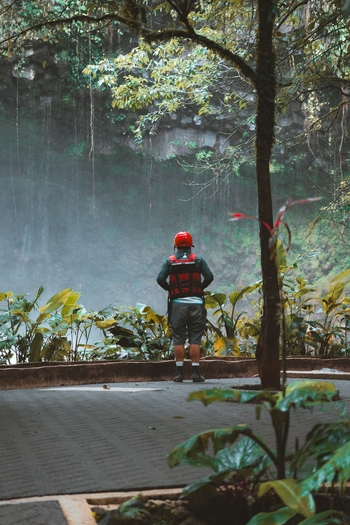 View of a person wearing a red helmet and life vest standing on a stone path in a lush tropical forest with a misty waterfall in the background in Costa Rica.