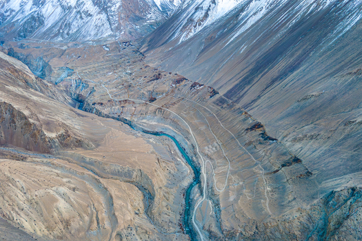 Aerial view of a winding river cutting through the rugged mountainous terrain of Chapursan Valley etched with pathways against the backdrop of snow capped peaks Hunza Nagar Gilgit Pakistan. Aerial view of a winding river cutting through the rugged mountainous terrain of Chapursan Valley etched with pathways against the backdrop of snow capped peaks Hunza Nagar Gilgit Pakistan.