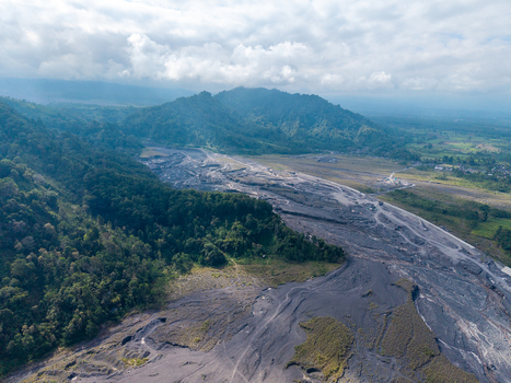 Aerial view of the lava path of Mount Semeru featuring dark volcanic deposits cutting through lush green forests under a cloudy sky Candipuro East Java Indonesia. Aerial view of the lava path of Mount Semeru featuring dark volcanic deposits cutting through lush green forests under a cloudy sky Candipuro East Java Indonesia.