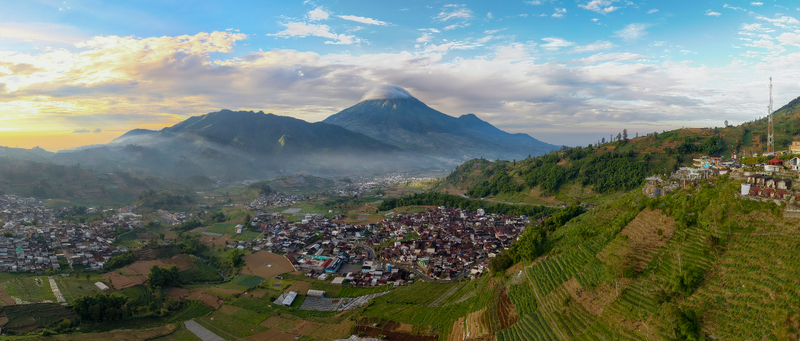 Aerial view of the misty Dieng plateau with Mount Sindoro volcano terraced fields and a dense village under a morning sky in Kecamatan Kejajar Jawa Tengah Indonesia.