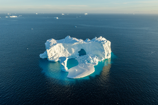 Aerial view of an arch iceberg floating in the deep blue waters of the Arctic Ocean surrounded by smaller ice fragments in Greenland. Aerial view of an arch iceberg floating in the deep blue waters of the Arctic Ocean surrounded by smaller ice fragments in Greenland.