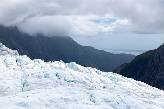 View of the icy glacier s textured surface meeting the distant dark forested mountains under a cloudy sky Franz Josef Glacier West Coast Region New Zealand. View of the icy glacier s textured surface meeting the distant dark forested mountains under a cloudy sky Franz Josef Glacier West Coast Region New Zealand.