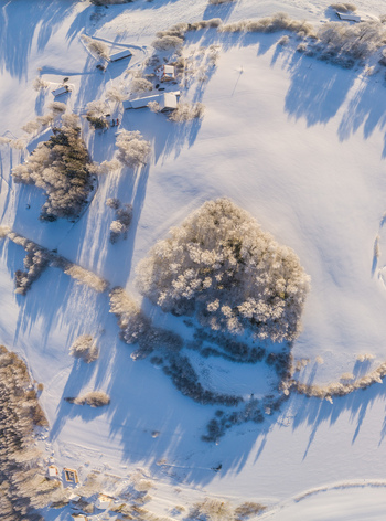 Aerial view of a snow covered rural landscape featuring frost covered trees small wooden buildings and long shadows cast by the winter sun Latvia. Aerial view of a snow covered rural landscape featuring frost covered trees small wooden buildings and long shadows cast by the winter sun Latvia.