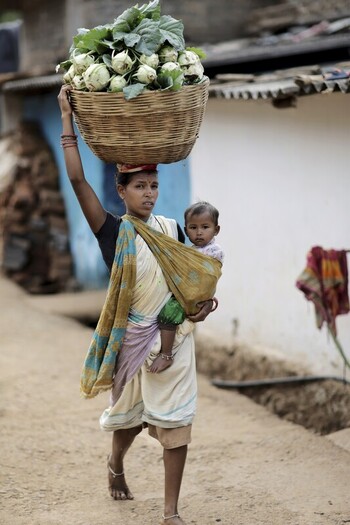 Odisha India 26 March 2026 View of a mother carries a basket of fresh produce atop her head her child nestled securely in a sling against the backdrop of rural homes. Odisha India 26 March 2026 View of a mother carries a basket of fresh produce atop her head her child nestled securely in a sling against the backdrop of rural homes.