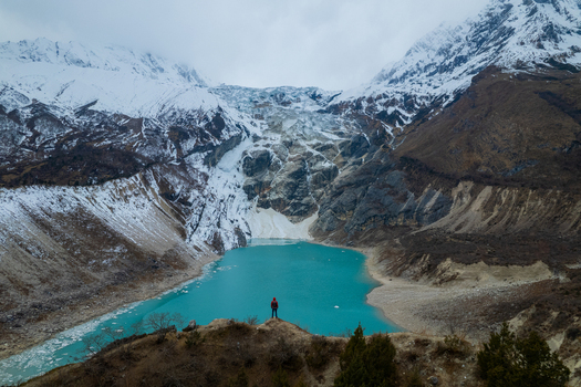 Aerial view of a person standing on a rocky ridge overlooking a turquoise glacial lake surrounded by snow capped Himalayan mountains in Nepal.