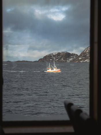 View of a fishing boat on the dark water under a cloudy sky framed by a window s edge creating a scene of solitude and maritime activity Brensholmen Troms Norway. View of a fishing boat on the dark water under a cloudy sky framed by a window s edge creating a scene of solitude and maritime activity Brensholmen Troms Norway.