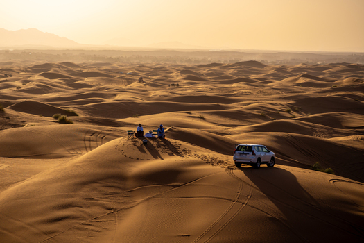 View of Erg Chebbi sand dunes with two people sitting on chairs and a white SUV parked in the desert landscape during golden hour in Errachidia Province Draa Tafilalet Morocco.