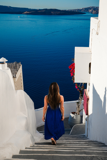 View of a woman in a blue dress walking down white stairs overlooking the deep blue Aegean Sea and volcanic caldera in Fira Greece. View of a woman in a blue dress walking down white stairs overlooking the deep blue Aegean Sea and volcanic caldera in Fira Greece.