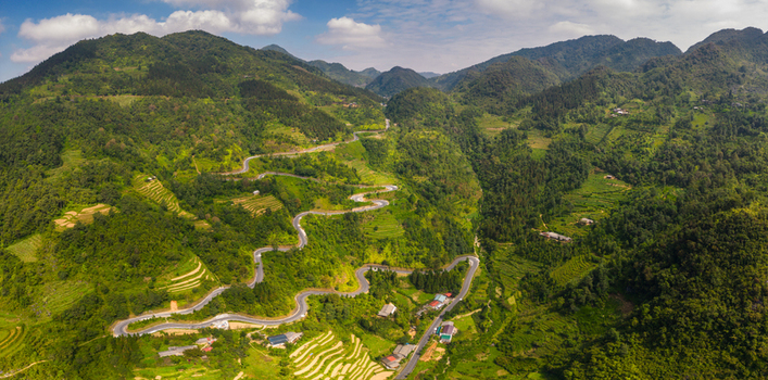 Aerial view of Chin Khoanh ramp winding through lush green mountains and terraced rice fields under a bright blue sky in Pho Bang Ha Giang Vietnam.