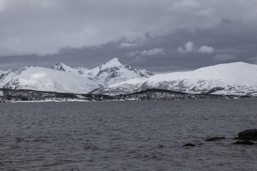 View of snow covered mountains and a calm fjord under a dramatic overcast sky in Tromso Troms Norway.