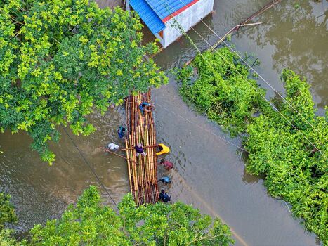 Banshkhali Upazila Bangladesh 10 July 2026 Aerial view of people navigating a bamboo raft through floodwaters surrounded by lush green trees and partially submerged buildings. Banshkhali Upazila Bangladesh 10 July 2026 Aerial view of people navigating a bamboo raft through floodwaters surrounded by lush green trees and partially submerged buildings.