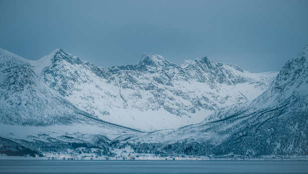 View of snow covered mountain peaks and a small coastal village with glowing lights at dusk in Sommaroy Troms Norway. View of snow covered mountain peaks and a small coastal village with glowing lights at dusk in Sommaroy Troms Norway.