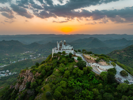 Aerial view of the historic Monsoon Palace perched on a hilltop surrounded by lush green hills under a dramatic sunset sky in Udaipur Rajasthan India.