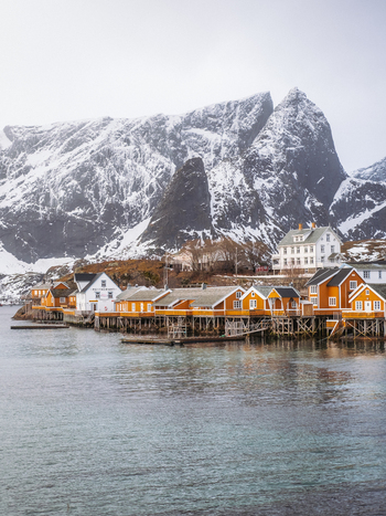 View of quaint yellow and white stilt houses nestled along the tranquil waters backed by snow dusted mountains piercing the sky Hamn&oslash;y Nordland Norway.