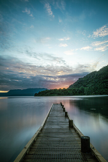 View of a wooden jetty extending into the calm waters of Lake Tarawera at sunset with distant hills and a soft cloudy sky in Rotorua Bay of Plenty Region New Zealand.