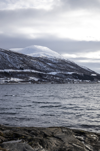 View of the snow covered mountains and coastal settlement across the choppy fjord waters under a cloudy sky in Tromso Troms Norway.