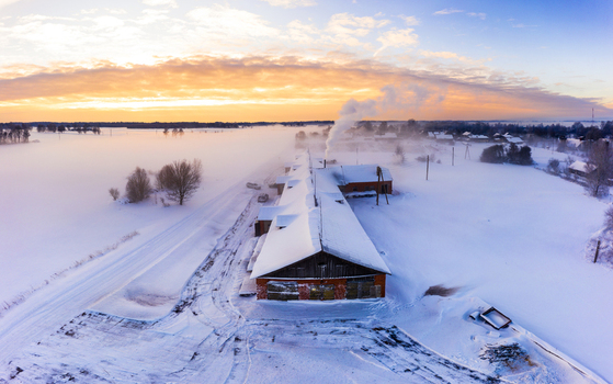 Aerial view of a rural building with smoke rising from a chimney in a snow covered landscape during a golden sunrise in Latvia. Aerial view of a rural building with smoke rising from a chimney in a snow covered landscape during a golden sunrise in Latvia.