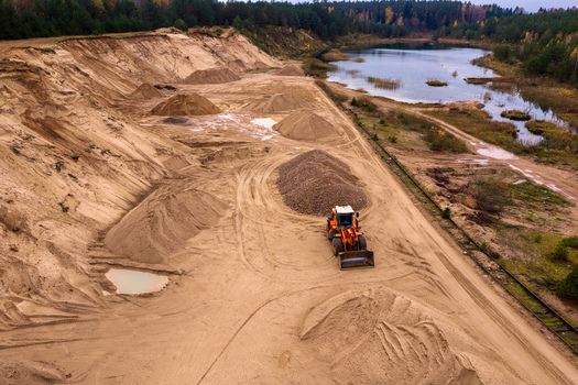 Aerial view of a sand quarry with an orange front loader working near piles of gravel and a calm lake surrounded by autumn forest in Nemencine Vilnius County Lithuania. Aerial view of a sand quarry with an orange front loader working near piles of gravel and a calm lake surrounded by autumn forest in Nemencine Vilnius County Lithuania.