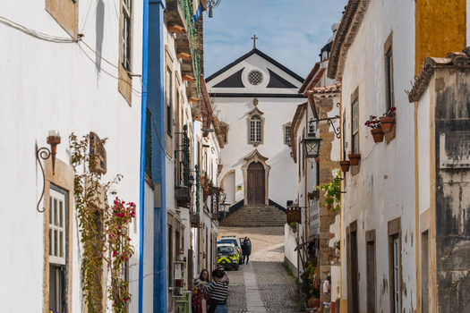 Obidos Portugal 13 May 2026 View of a narrow cobblestone street in the historic town of Obidos featuring traditional white washed buildings flower pots and a church at the end of the path. Obidos Portugal 13 May 2026 View of a narrow cobblestone street in the historic town of Obidos featuring traditional white washed buildings flower pots and a church at the end of the path.