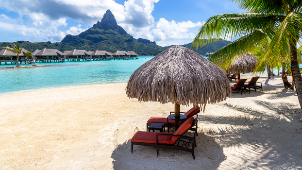 View of overwater bungalows on the turquoise lagoon with Mount Otemanu and sun loungers under a thatched umbrella on the sandy beach in Bora Bora Leeward Islands French Polynesia.