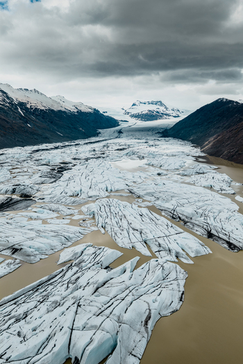 Aerial view of Vatnajokull glacier featuring expansive ice formations and glacial meltwater pools surrounded by rugged mountains under a cloudy sky in Iceland.