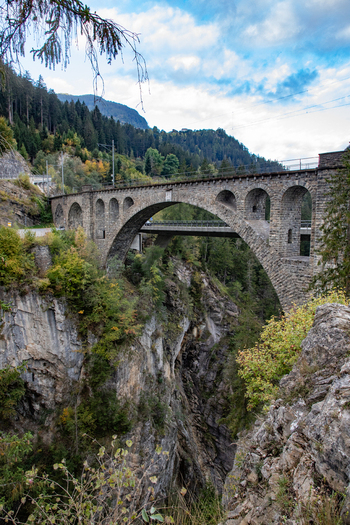 View of a historic stone arch bridge spanning a deep gorge in the Swiss Alps surrounded by lush pine forests and rugged mountain terrain under a cloudy sky in Airolo Ticino Switzerland. View of a historic stone arch bridge spanning a deep gorge in the Swiss Alps surrounded by lush pine forests and rugged mountain terrain under a cloudy sky in Airolo Ticino Switzerland.