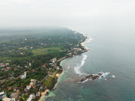 Aerial view of the tropical coastline with turquoise waters rocky outcrops and lush green palm trees under a misty sky in Unawatuna Southern Province Sri Lanka.
