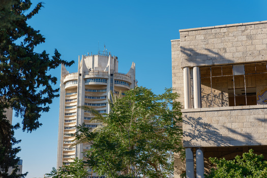View of the Le Royal Hotel tower rising behind a historic stone building with palm trees under a clear blue sky in Amman Amman Governorate Jordan.