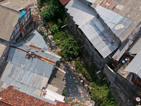 Aerial view of a narrow polluted canal flowing between dense residential buildings with corrugated metal roofs in Kecamatan Denpasar Barat Bali Indonesia. Aerial view of a narrow polluted canal flowing between dense residential buildings with corrugated metal roofs in Kecamatan Denpasar Barat Bali Indonesia.