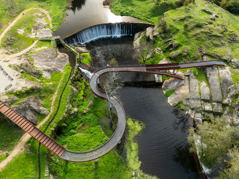 Aerial view of Acude da Ribeira weir featuring a modern sinuous metal walkway over the cascading water and green rocky riverbanks in Ervedal Coimbra Portugal. Aerial view of Acude da Ribeira weir featuring a modern sinuous metal walkway over the cascading water and green rocky riverbanks in Ervedal Coimbra Portugal.