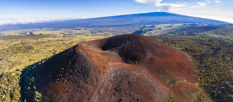 Aerial view of the red volcanic cinder cone of Mauna Kea surrounded by lava fields and sparse vegetation under a clear blue sky in Hawaii County Hawaii United States.