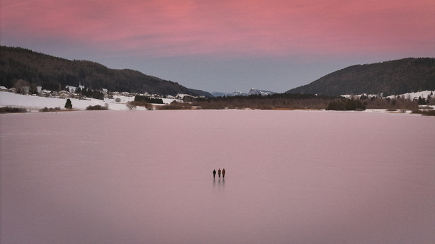 Aerial view of three figures stand silhouetted on the vast frozen expanse of the lake under the soft pink hues of the winter sky Les Rousses Bourgogne Franche Comté France. Aerial view of three figures stand silhouetted on the vast frozen expanse of the lake under the soft pink hues of the winter sky Les Rousses Bourgogne Franche Comté France.