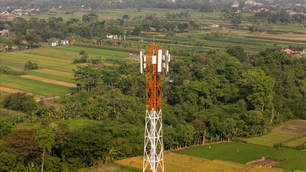 Aerial view of a telecommunications tower with an orange and white lattice structure standing amidst lush green agricultural fields and rural landscape in Indonesia.