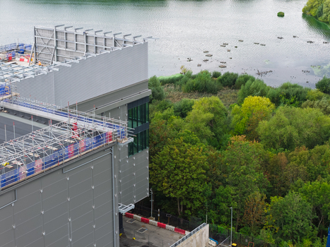Aerial view of a modern data center building featuring rooftop scaffolding and metallic cladding situated next to a tranquil lake and lush green woodland in the United Kingdom.
