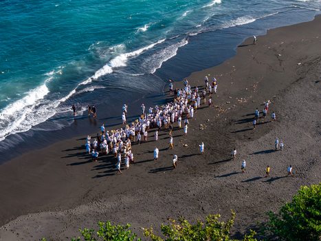 Aerial view of Ngelarung Sea Offering Ceremony at Pura Segara Goa Lawah with people in white clothing gathered on the black sand beach by the turquoise sea in Dawan Bali Indonesia.