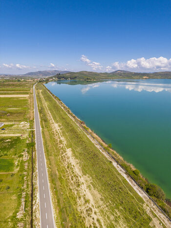 Aerial view of a road running alongside Liqeni Thanes turquoise lake bordered by green fields and rolling hills in Lushnje Fier County Albania. Aerial view of a road running alongside Liqeni Thanes turquoise lake bordered by green fields and rolling hills in Lushnje Fier County Albania.