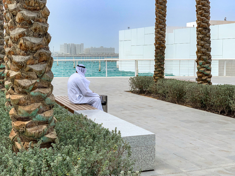 View of a man in traditional Emirati clothing sitting on a bench along a waterfront promenade with palm trees and modern buildings in Dubai Dubai United Arab Emirates. View of a man in traditional Emirati clothing sitting on a bench along a waterfront promenade with palm trees and modern buildings in Dubai Dubai United Arab Emirates.