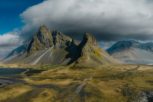 Aerial view of the dramatic Vestrahorn mountain range at Stokksnes featuring jagged peaks dark volcanic sand and a winding coastal road under a moody cloud filled sky in Iceland.