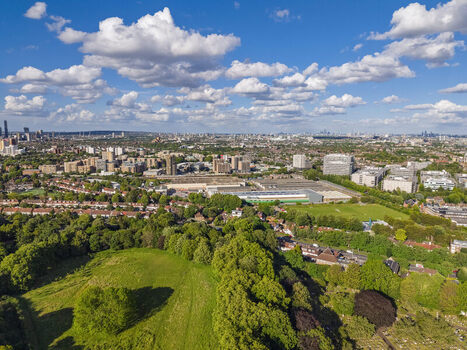 Aerial view of Gunnersbury Park with lush green trees and open fields overlooking the urban landscape and residential buildings of the city under a bright blue sky with scattered clouds London England United Kingdom. Aerial view of Gunnersbury Park with lush green trees and open fields overlooking the urban landscape and residential buildings of the city under a bright blue sky with scattered clouds London England United Kingdom.