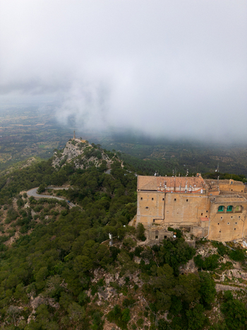 Aerial view of the Sanctuary of Sant Salvador on a misty hilltop surrounded by lush green forests and winding roads in Felanitx Illes Balears Spain. Aerial view of the Sanctuary of Sant Salvador on a misty hilltop surrounded by lush green forests and winding roads in Felanitx Illes Balears Spain.
