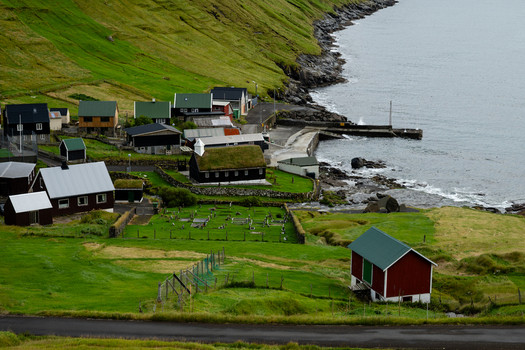 View of a small coastal village with traditional grass roofed houses a historic church and a stone pier along the rugged green coastline of the Faroe Islands. View of a small coastal village with traditional grass roofed houses a historic church and a stone pier along the rugged green coastline of the Faroe Islands.
