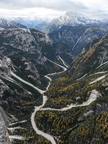 Aerial view of a winding river cutting through the heart of Rienza valley framed by rugged mountains with snow dusted peaks creating a dramatic contrast South Tyrol Italy.