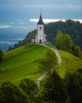 Aerial view of the Church of Saints Primus and Felician perched on a lush green hill with a winding path and misty mountains in the background in Jamnik Kranj Slovenia.