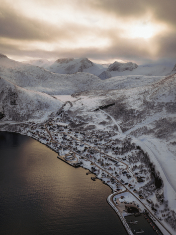Aerial view of a snow dusted village nestled between dark fjords and towering frosted mountains under a somber cloud streaked sky Fjordgård Troms Norway. Aerial view of a snow dusted village nestled between dark fjords and towering frosted mountains under a somber cloud streaked sky Fjordgård Troms Norway.