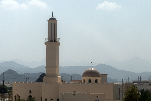 View of a mosque featuring a tall minaret and a central dome set against a backdrop of rugged mountains in Muscat Muscat Governorate Oman. View of a mosque featuring a tall minaret and a central dome set against a backdrop of rugged mountains in Muscat Muscat Governorate Oman.