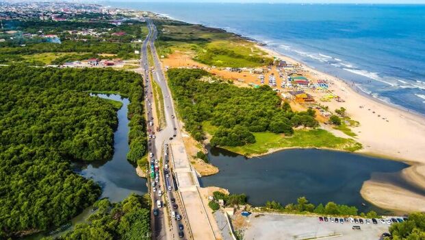 Aerial view of a coastal highway running alongside lush mangrove forests and a sandy beach with parked cars and ocean waves in Ghana. Aerial view of a coastal highway running alongside lush mangrove forests and a sandy beach with parked cars and ocean waves in Ghana.
