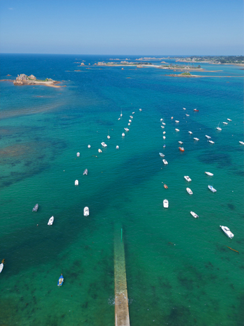 Aerial view of Plage de Port Blanc with numerous white boats anchored in turquoise waters near a stone pier and rocky islands under a clear blue sky in Penvenan Bretagne France. Aerial view of Plage de Port Blanc with numerous white boats anchored in turquoise waters near a stone pier and rocky islands under a clear blue sky in Penvenan Bretagne France.