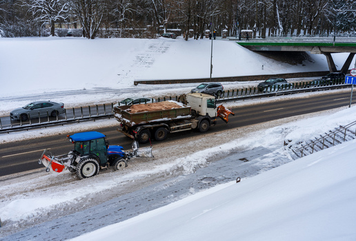 View of a blue tractor with a snow plow and a dump truck clearing snow from a road during winter in Vilnius Vilnius County Lithuania. View of a blue tractor with a snow plow and a dump truck clearing snow from a road during winter in Vilnius Vilnius County Lithuania.