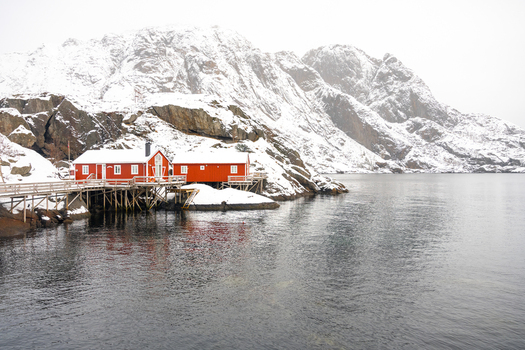 View of traditional red wooden cabins on stilts over the water with snow covered mountains in the background in Leknes Lofoten Norway.